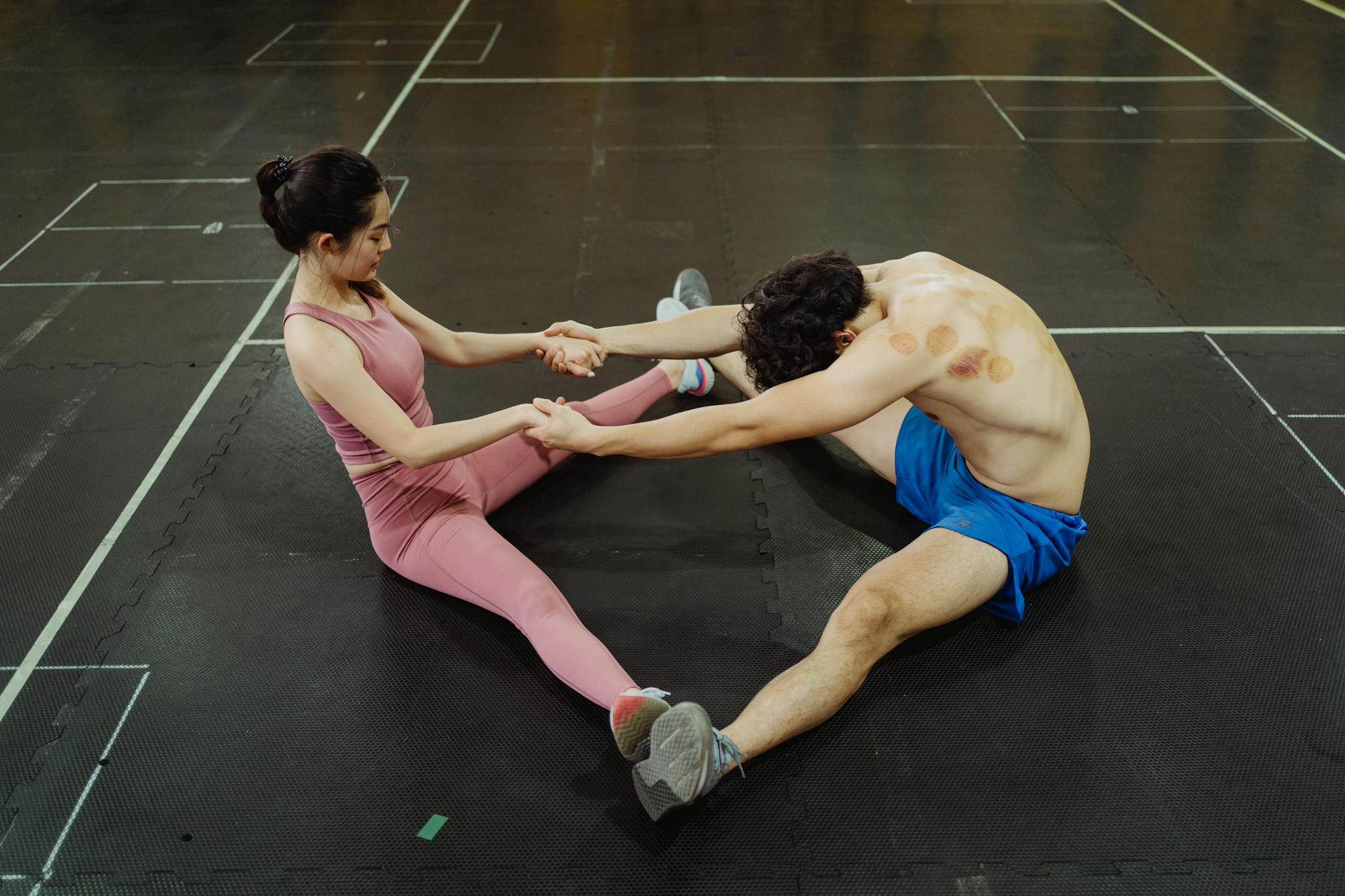 A man and woman performing partner stretching exercises on a gym floor, enhancing flexibility and teamwork.