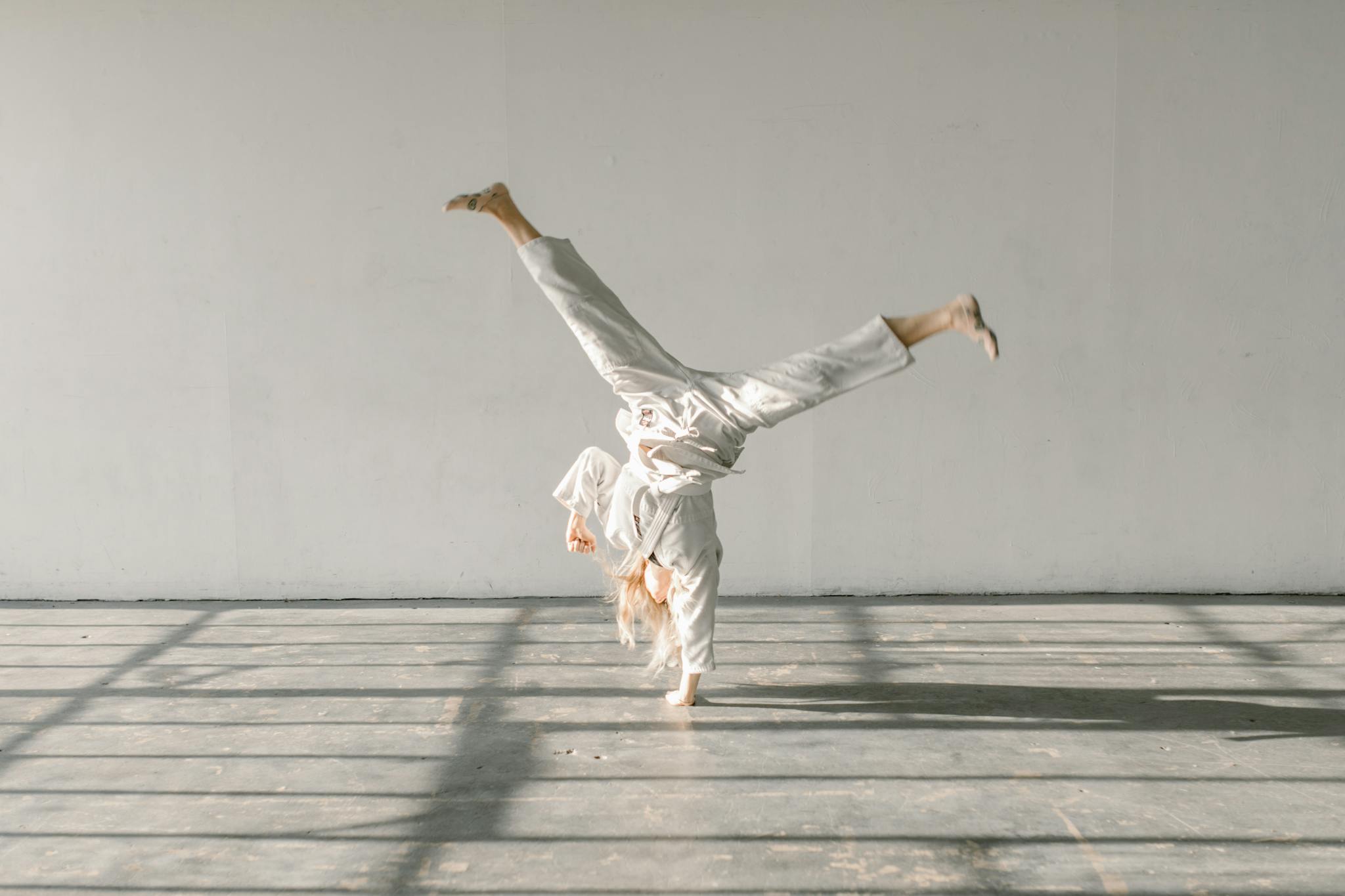 A woman in a white gi performing a handstand in a well-lit gym setting.